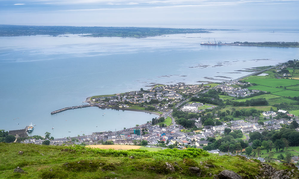 A stunning aerial view beautifully showcases a picturesque coastal Carlingford town, surrounded by vibrant greenery and calm waters, all under an expansive, open sky above, Ireland
